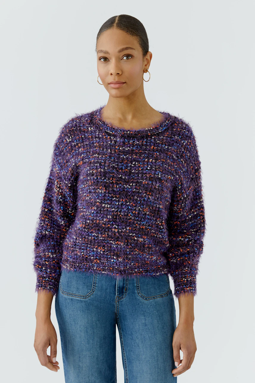 Frau im lila Pullover mit grobem Strickmuster, kombiniert mit blauen Jeans, schaut direkt in die Kamera.