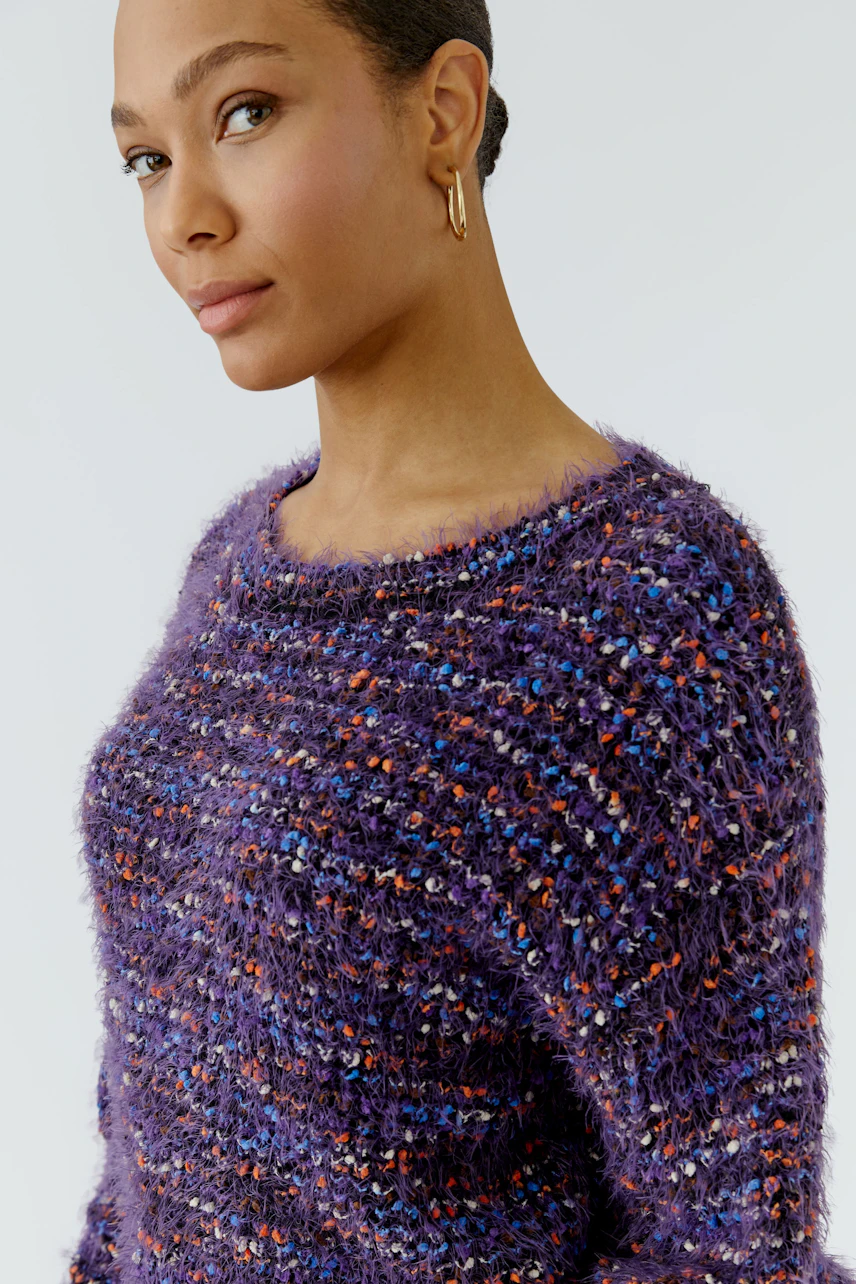 Frau im lila Pullover mit bunter Struktur, lässig und modern. Blickt selbstbewusst zur Seite.