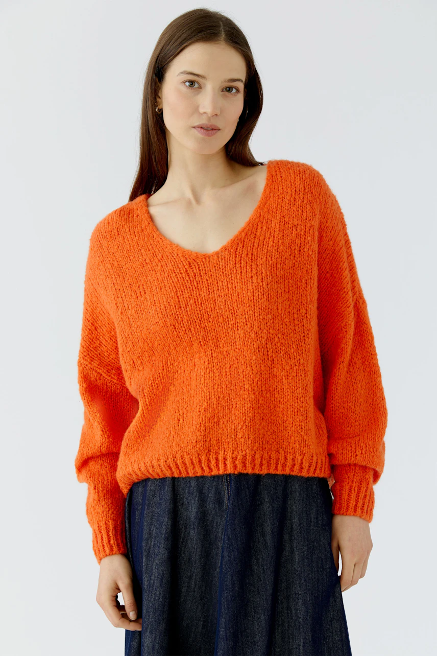 Frau im leuchtend orangefarbenen Pullover mit V-Ausschnitt, kombiniert mit einem dunklen Rock.