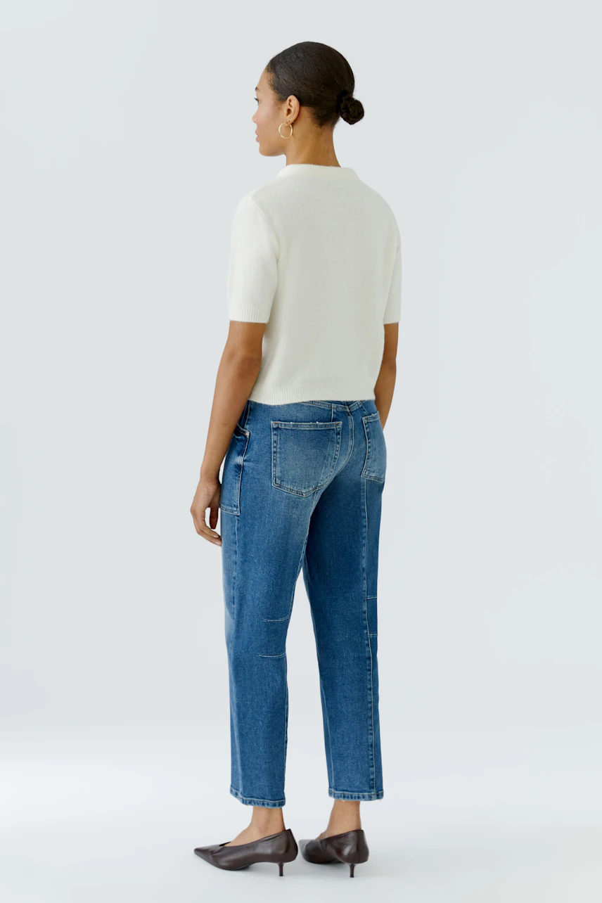 Frau im weißen Pullover und blauer Jeans, seitlich stehend, zeigt lässigen Look.