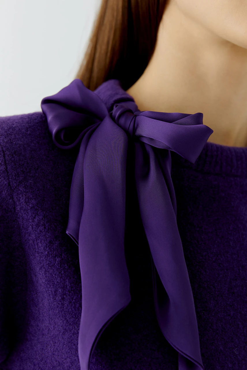 Woman in a purple pullover with a large bow detail on the shoulder, showcasing a stylish and bold look.