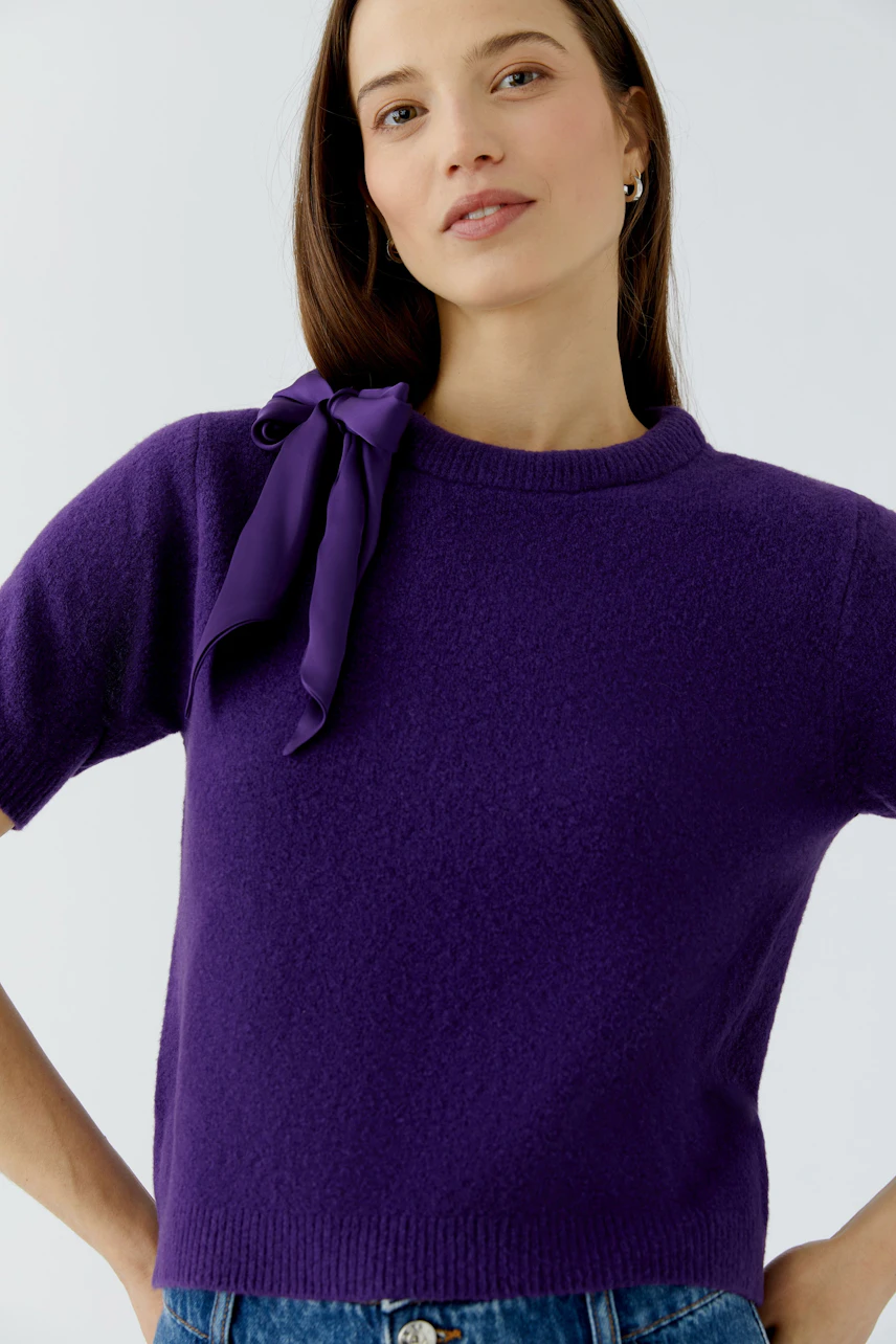 Woman in a purple pullover with a bow detail, short sleeves, and casual style.