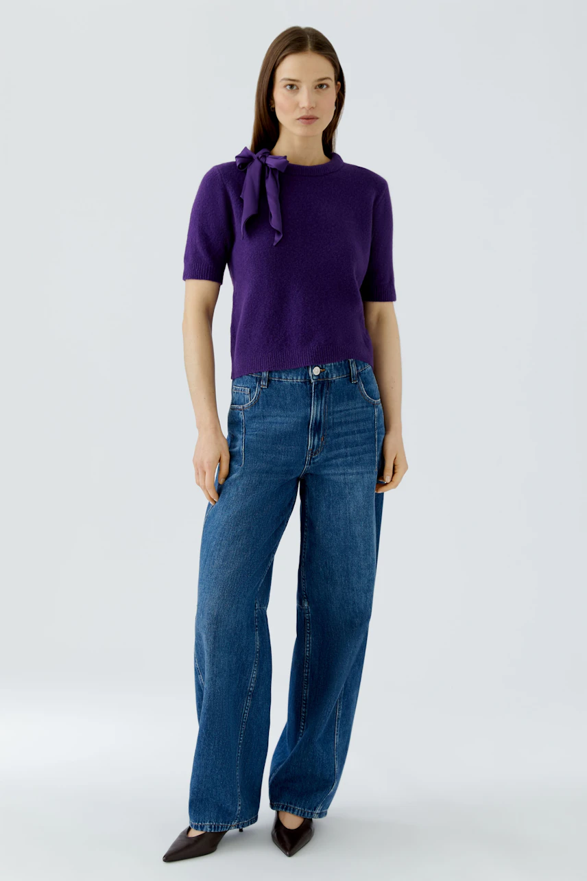Casual women's pullover: Woman in purple pullover with bow detail, paired with blue jeans, stands confidently.