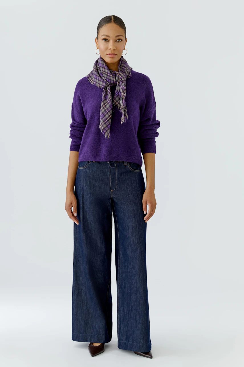 Casual scarf: Woman in a purple pullover and wide jeans, wearing a plaid scarf, standing confidently.