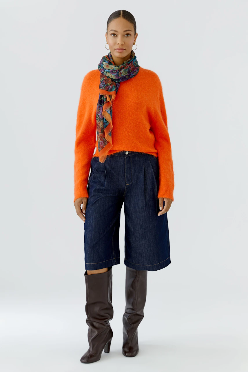 Colorful scarf: Woman in orange pullover, denim culottes, and brown boots, wearing a vibrant patterned scarf.