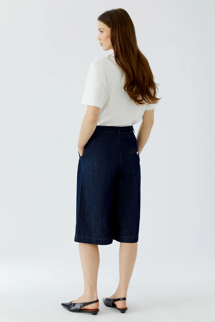 Frau in weißem Shirt und Bundfaltenhose aus Denim, seitlich stehend, mit Händen in den Taschen.