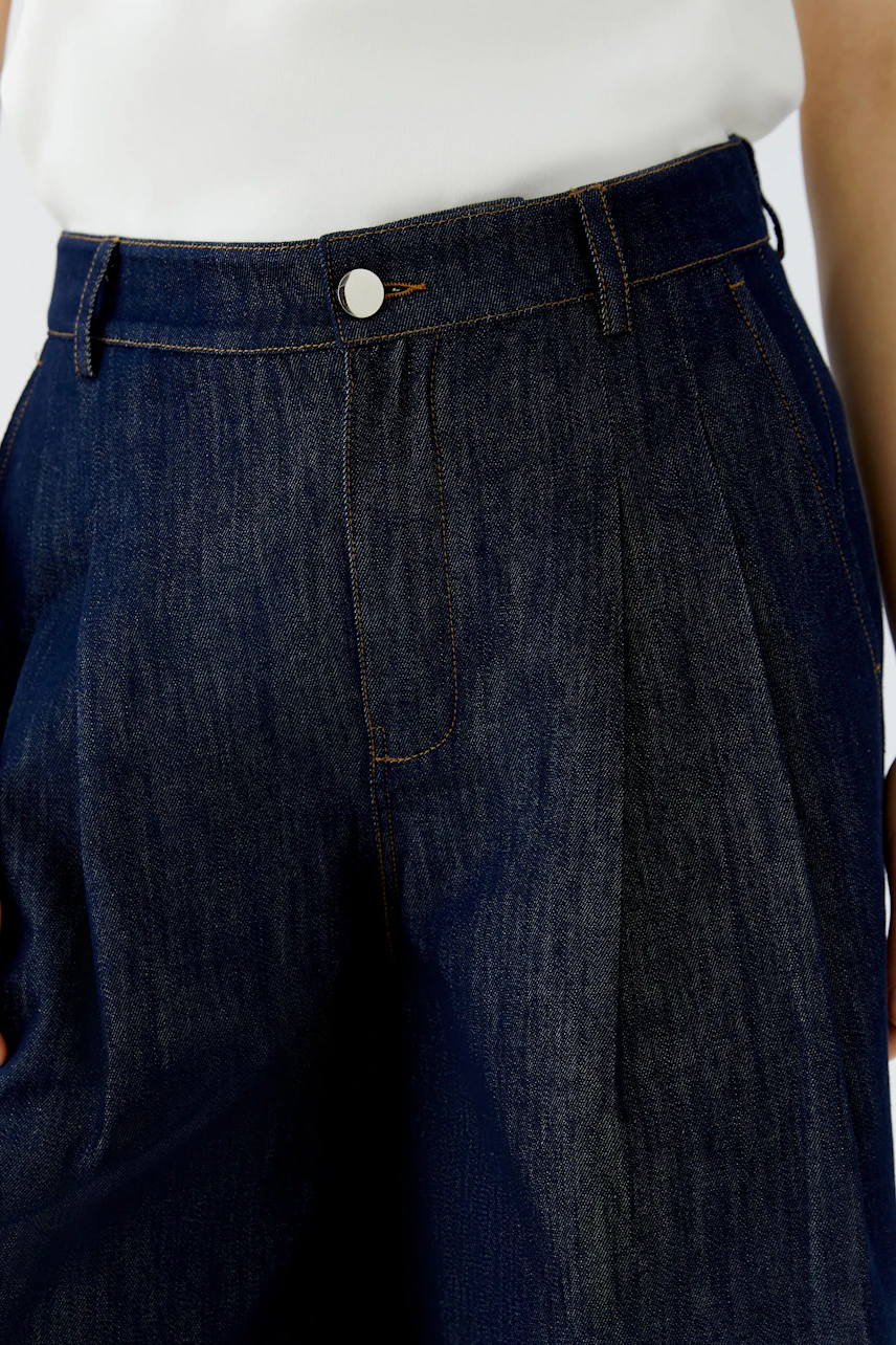 Damen-Bundfaltenhose: Frau in dunkelblauer Hose aus Denim, kombiniert mit weißem Oberteil, zeigt modernen Stil.