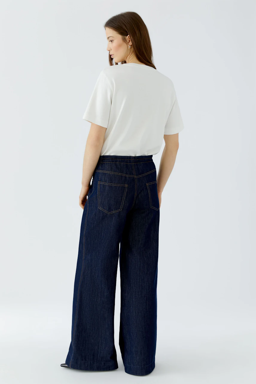 Jeans Wide Leg: Frau in dunkelblauer, weit geschnittener Jeans und weißem T-Shirt von hinten.