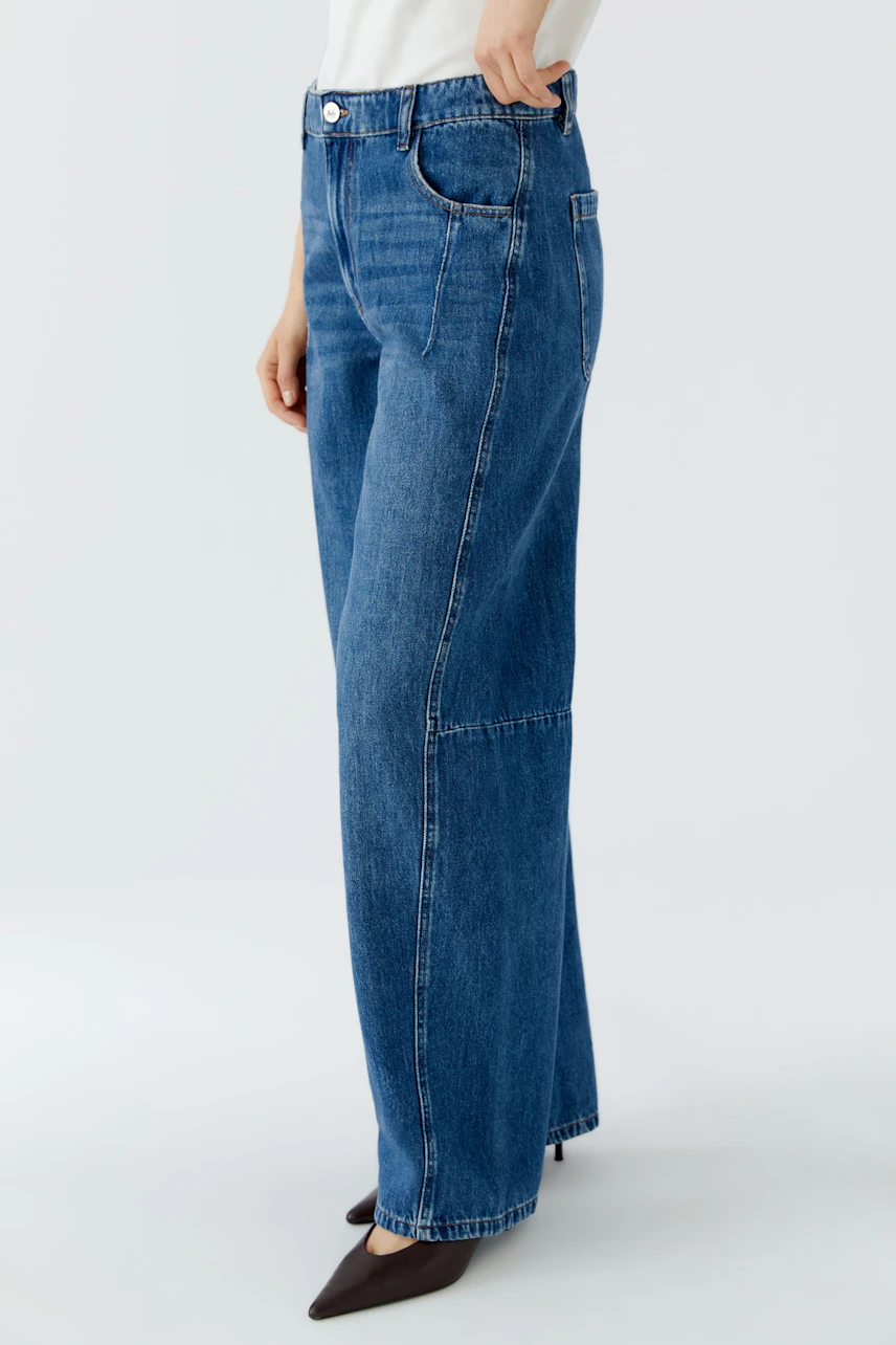 Jeans Barrel: Frau in blauer, weit geschnittener Jeans mit weißem Oberteil, seitlich stehend.