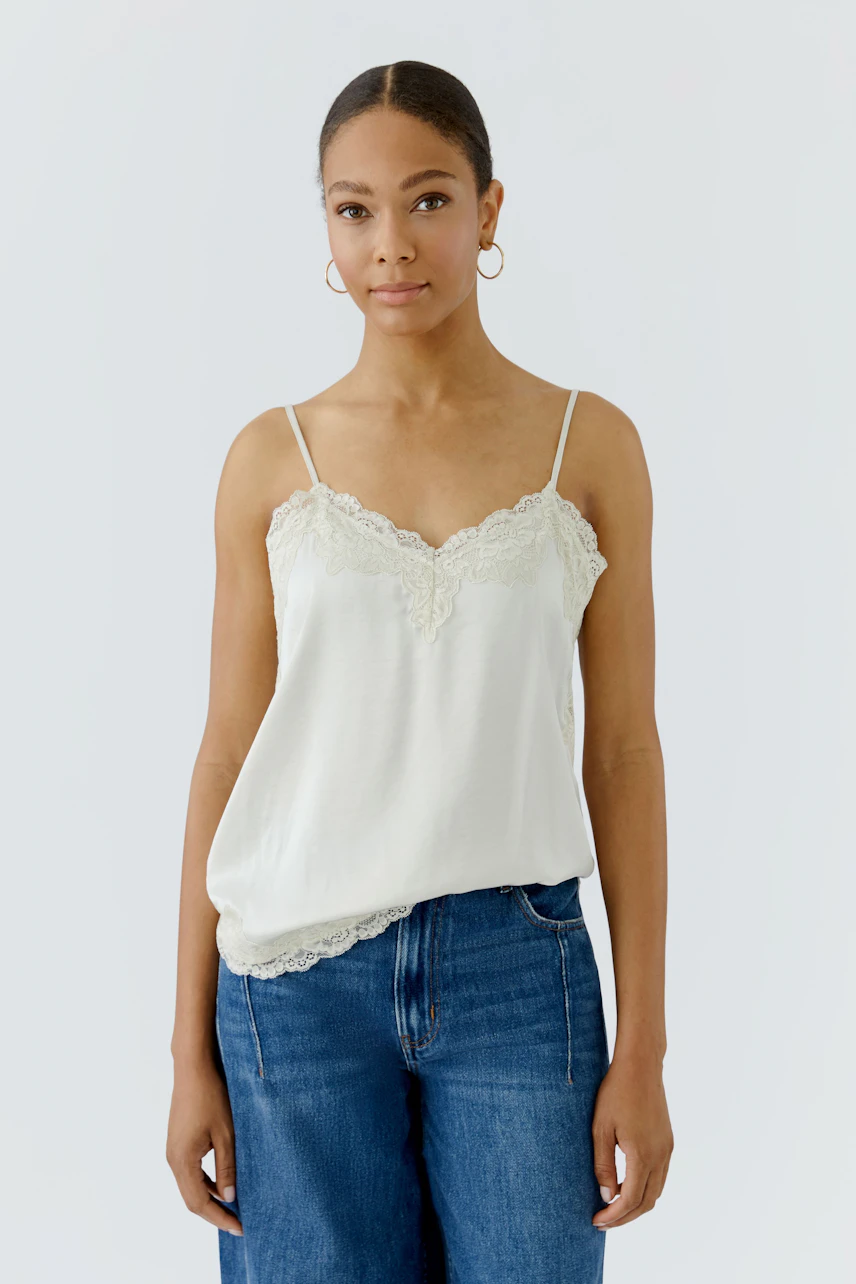 Damen-Top: Frau im weißen Spitzentop mit schmalen Trägern und blauer Jeans, schlicht und stilvoll.