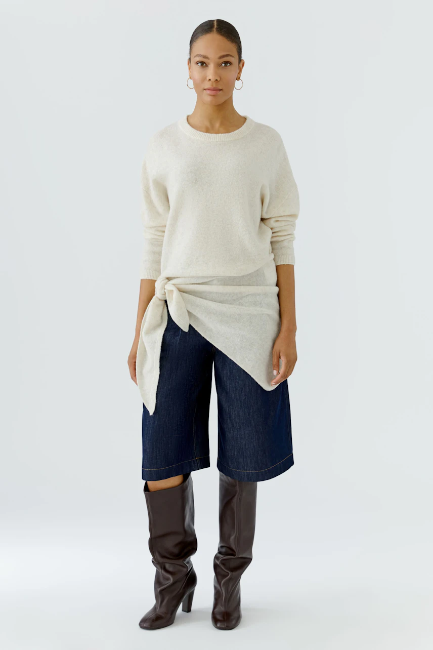 Woman in cream pullover with tied scarf, denim culottes, and brown boots, standing confidently.