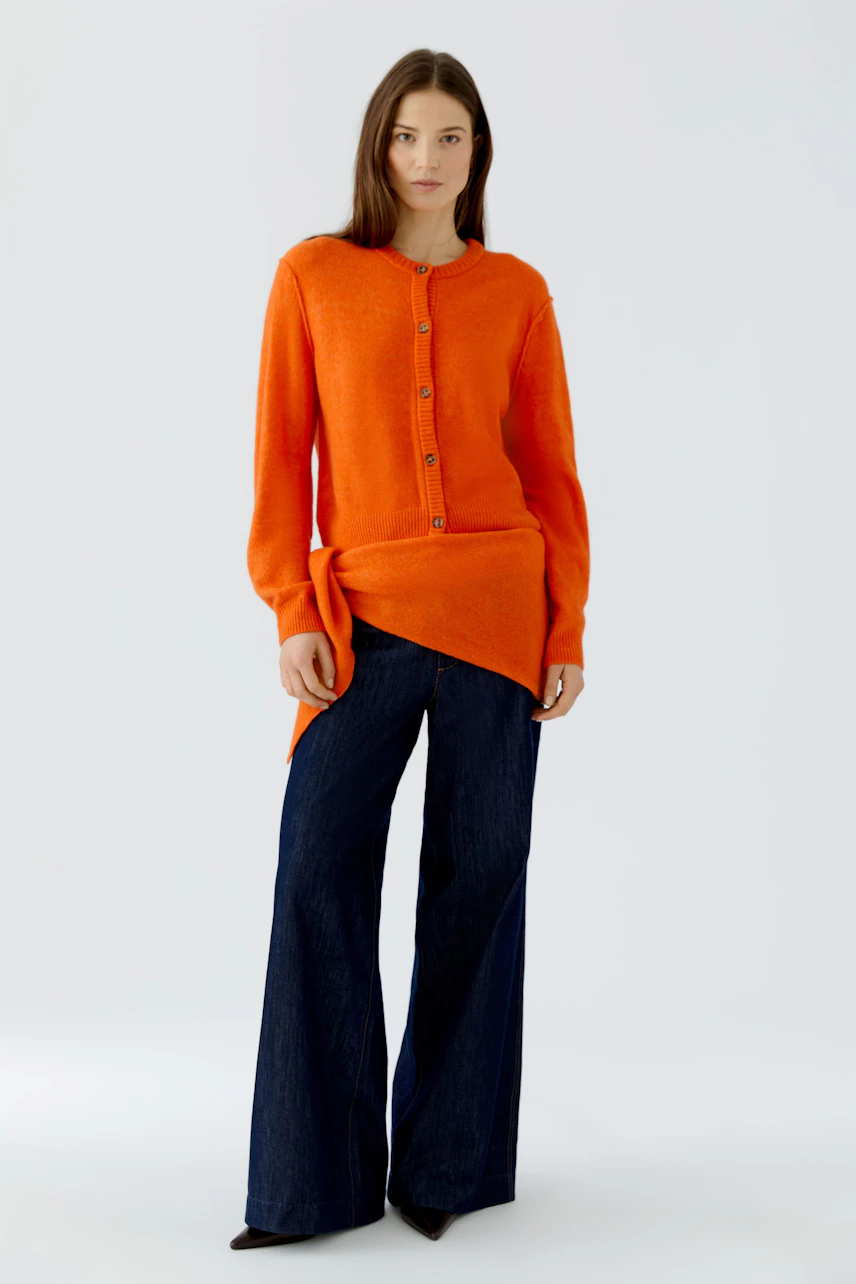 Woman in orange cardigan and wide-leg jeans, holding a scarf. Casual and stylish look.