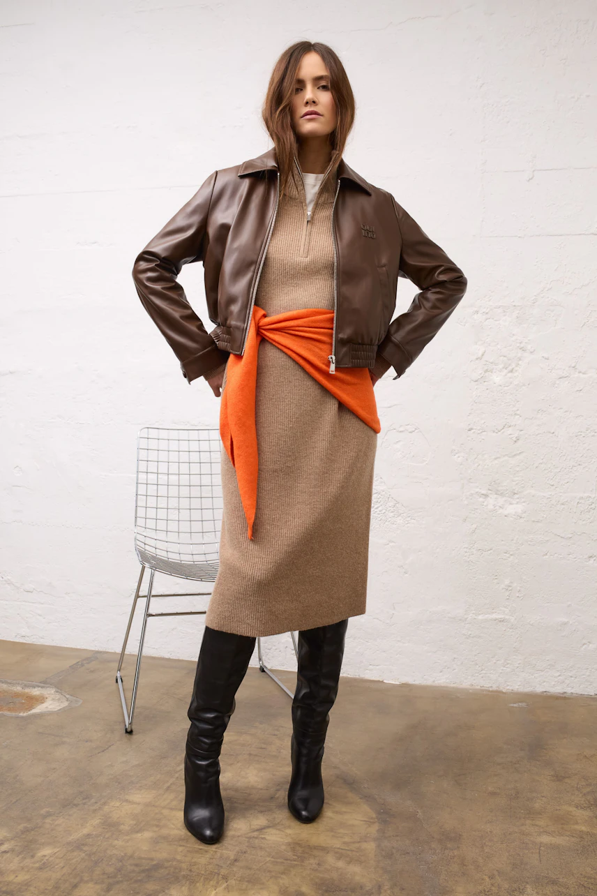 Woman in brown dress with orange scarf, leather jacket, and black boots stands confidently.