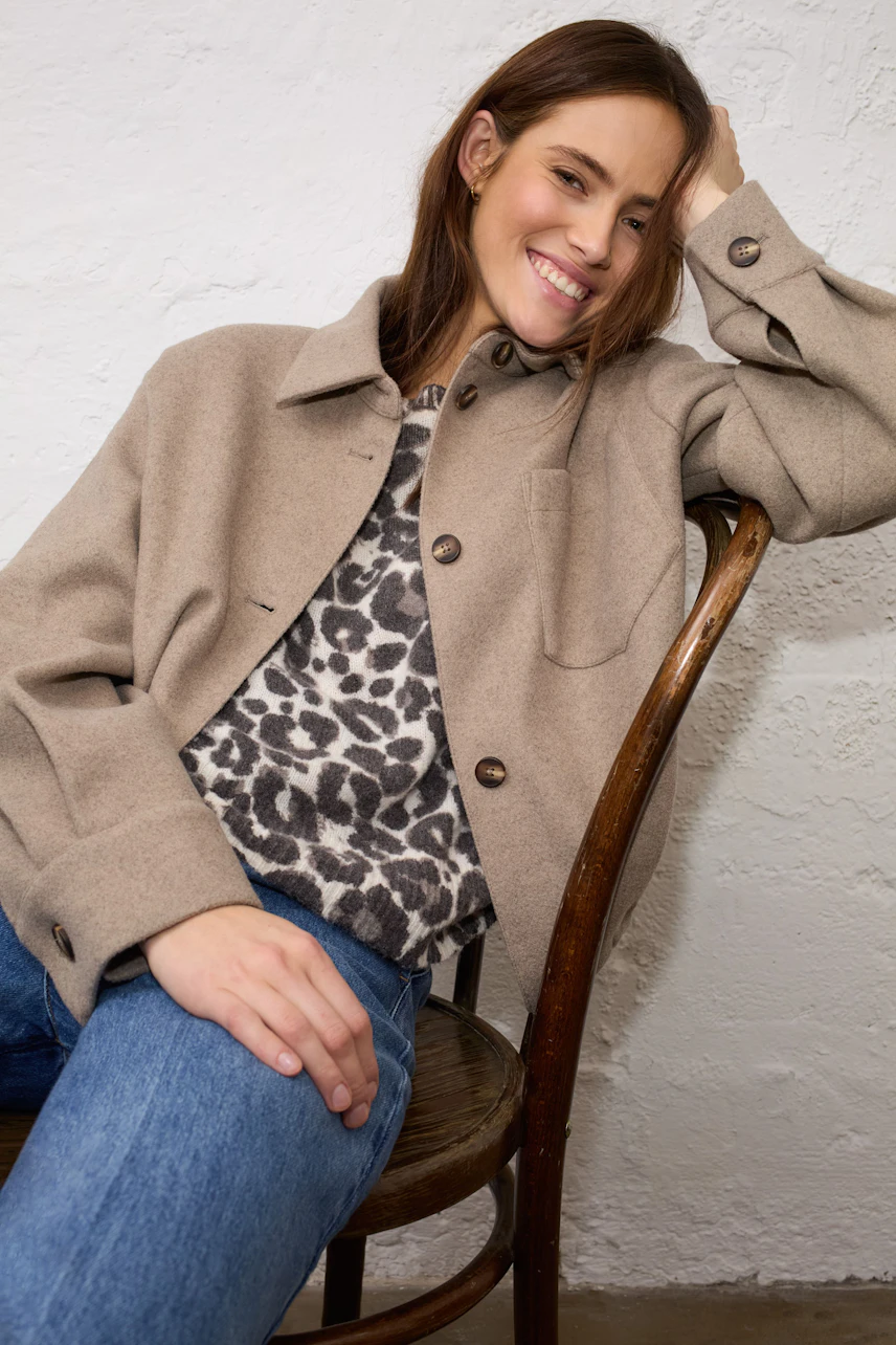 Casual women's jacket: Woman in beige jacket over leopard print top, sitting relaxed on a chair.