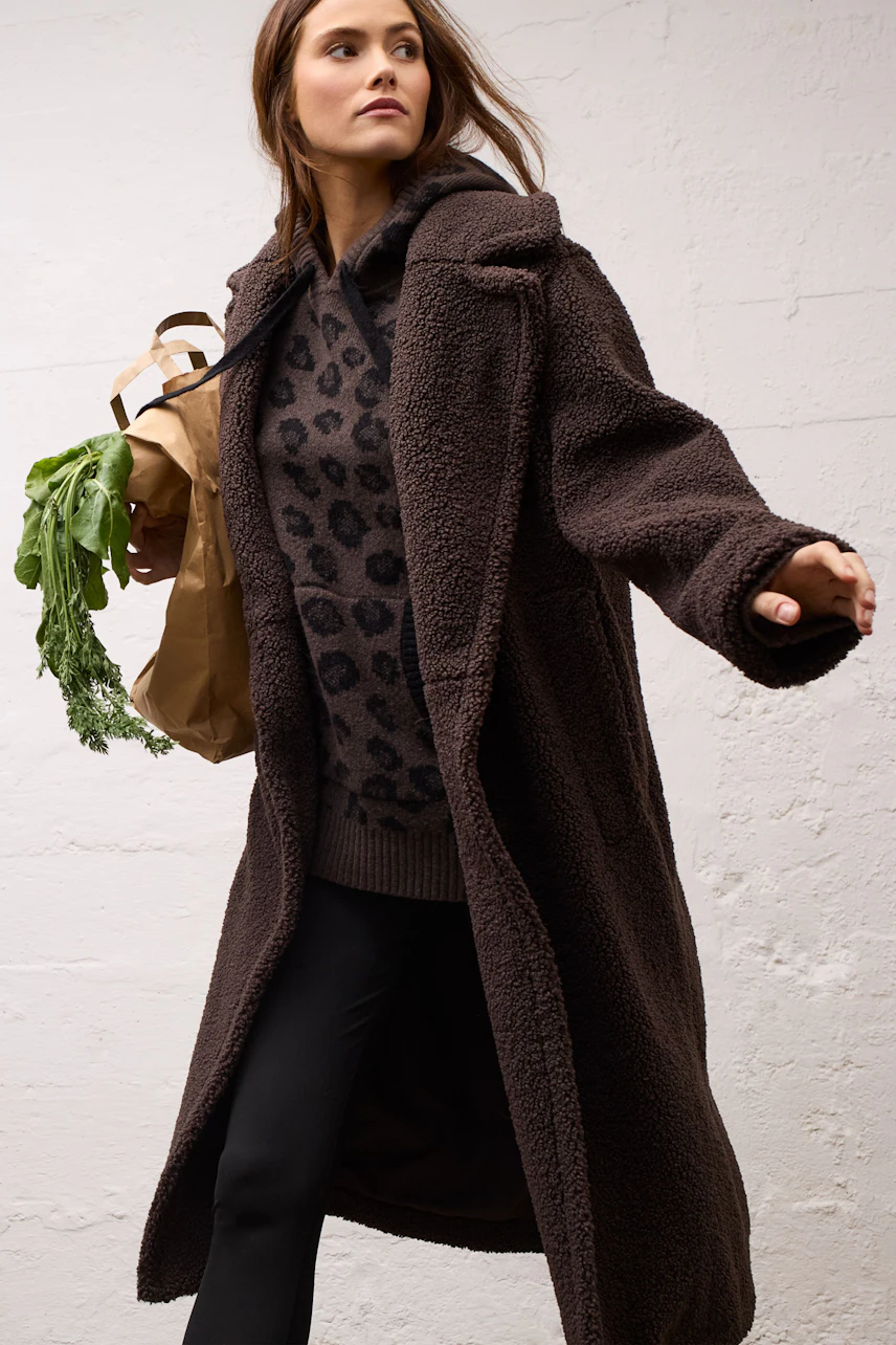 Woman in a cozy leopard print pullover and long brown coat, holding groceries, walking confidently.