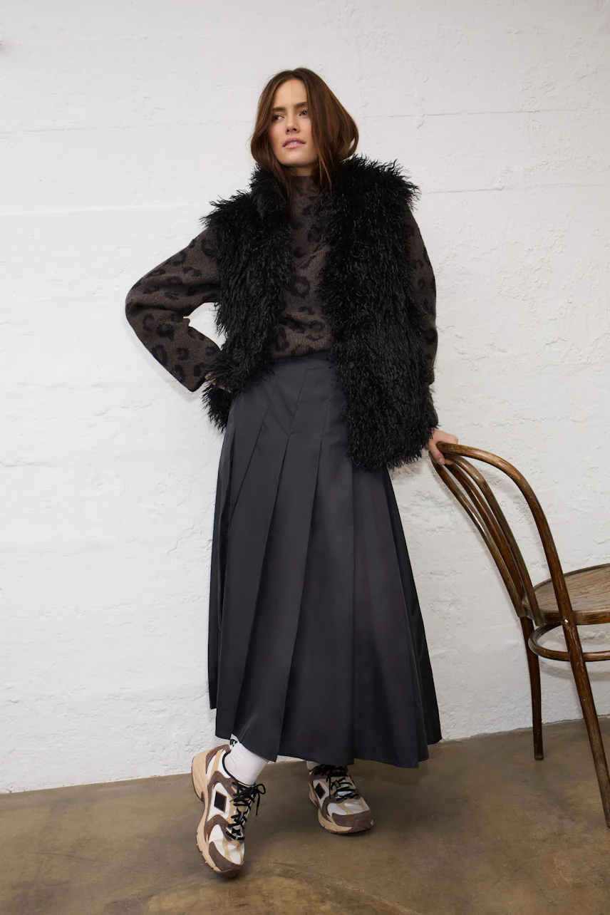 Woman in a patterned pullover with a black skirt and fluffy vest, standing confidently.