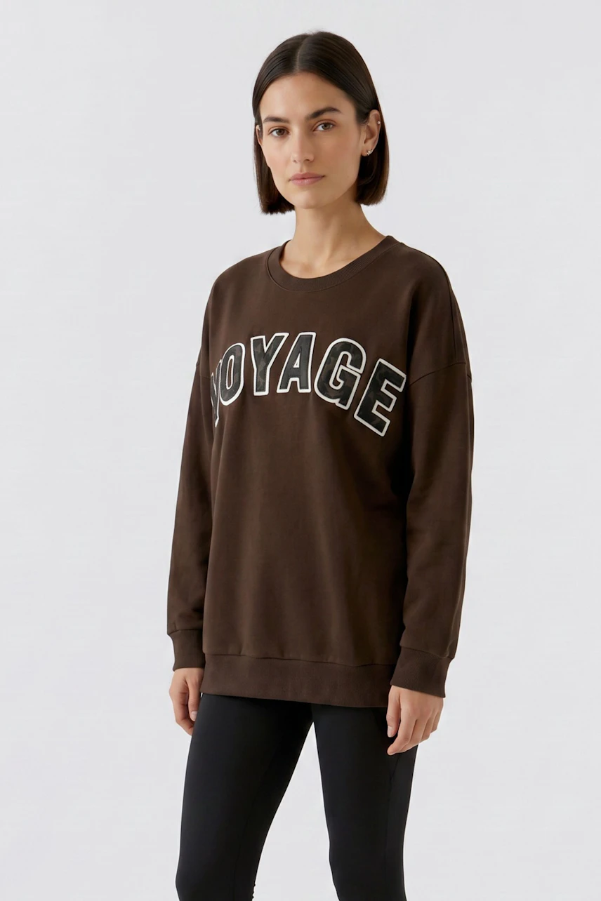 Junge Frau im braunen Sweatshirt mit "VOYAGE"-Aufdruck, lässig kombiniert mit passender Hose.