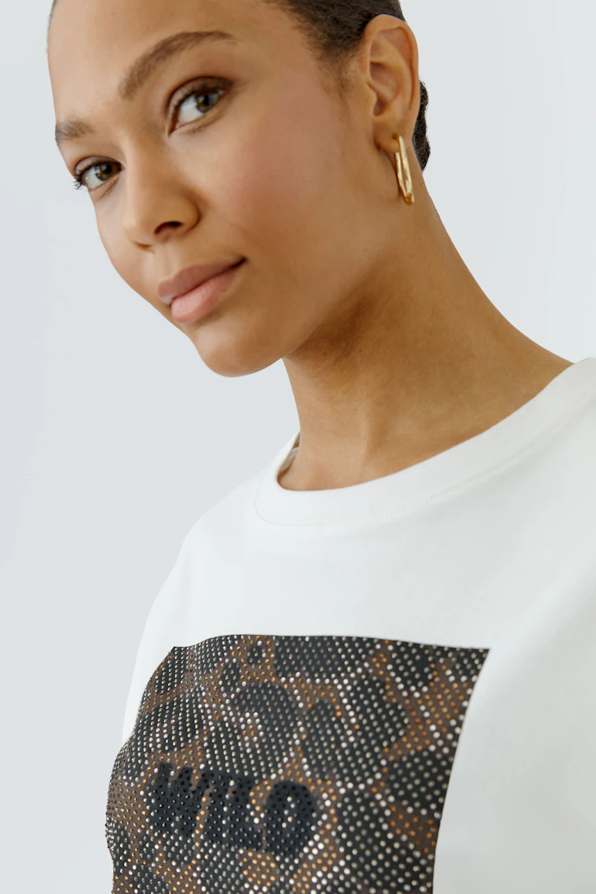 Damen-T-Shirt: Frau im weißen T-Shirt mit auffälligem Print und goldenen Ohrringen schaut freundlich.