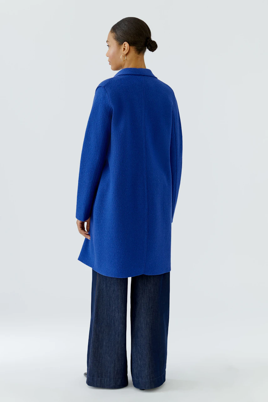 Frau im blauen Wollmantel, kombiniert mit dunkler Hose, von hinten zu sehen.