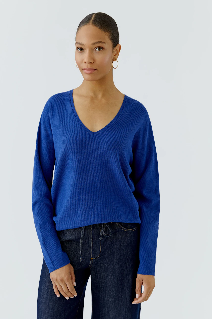 Casual women's pullover: Woman in a blue V-neck pullover and dark jeans, standing confidently.