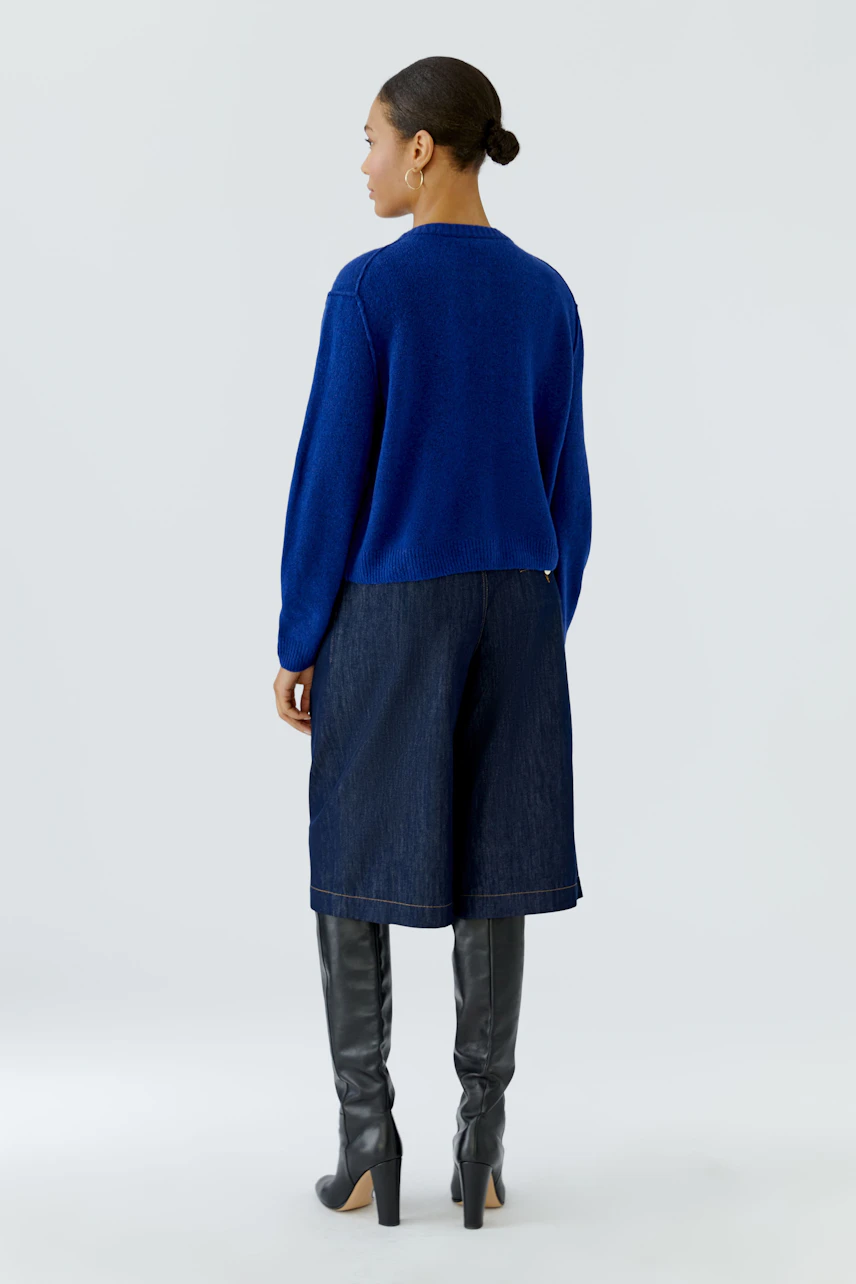 Blue knit cardigan: Woman in a blue cardigan, denim culottes, and black knee-high boots, facing away.