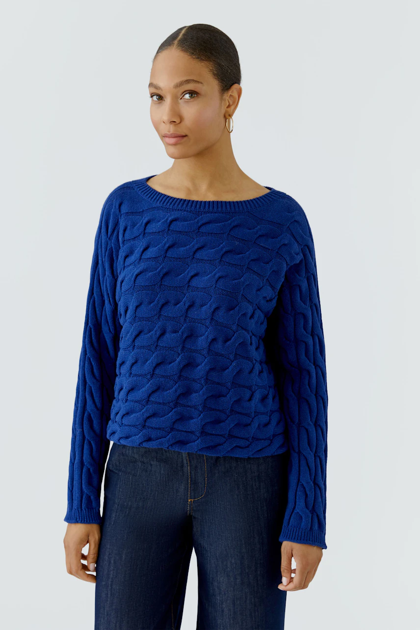 Frau im blauen Pullover mit Zopfmuster, kombiniert mit dunkler Jeans, schaut selbstbewusst.