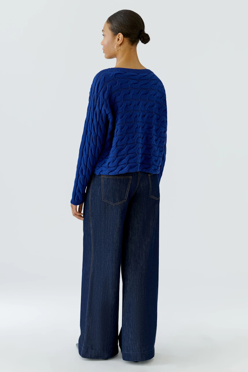 Frau im blauen Pullover mit Zopfmuster, kombiniert mit weiter Jeans. Rückansicht.