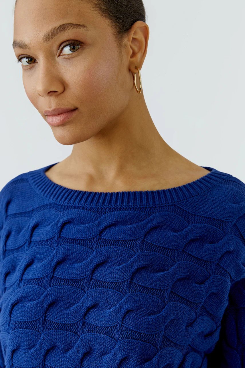 Frau im blauen Pullover mit Zopfmuster, goldene Ohrringe, lässiger Look.