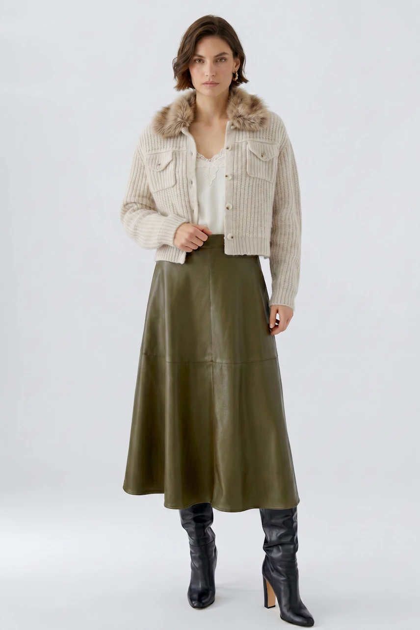 Woman in olive A-line skirt, cream jacket, and black boots, standing confidently.