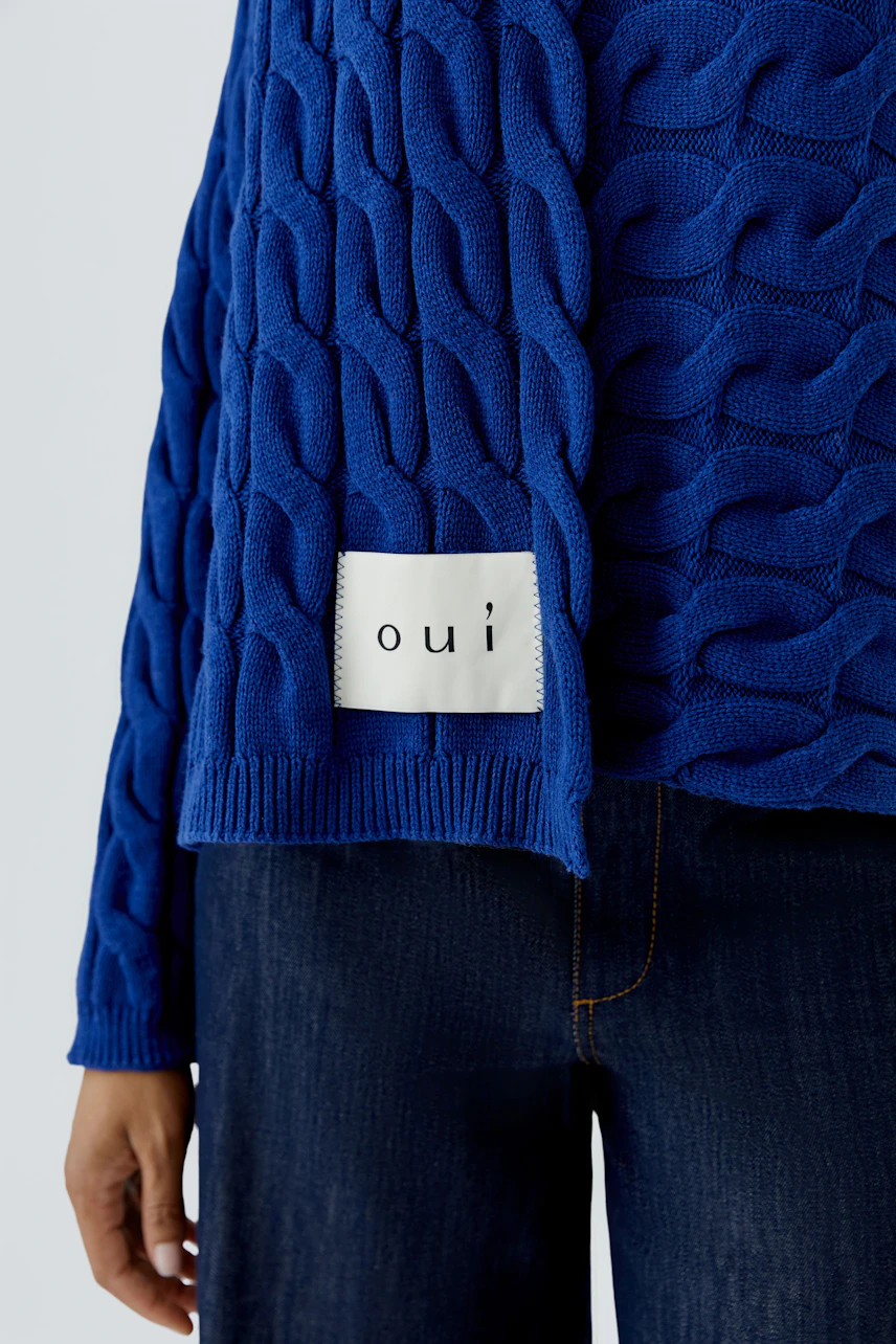 Blauer Strickpullover: Frau trägt Pullover mit Zopfmuster und Oui-Label, kombiniert mit dunkler Jeans.