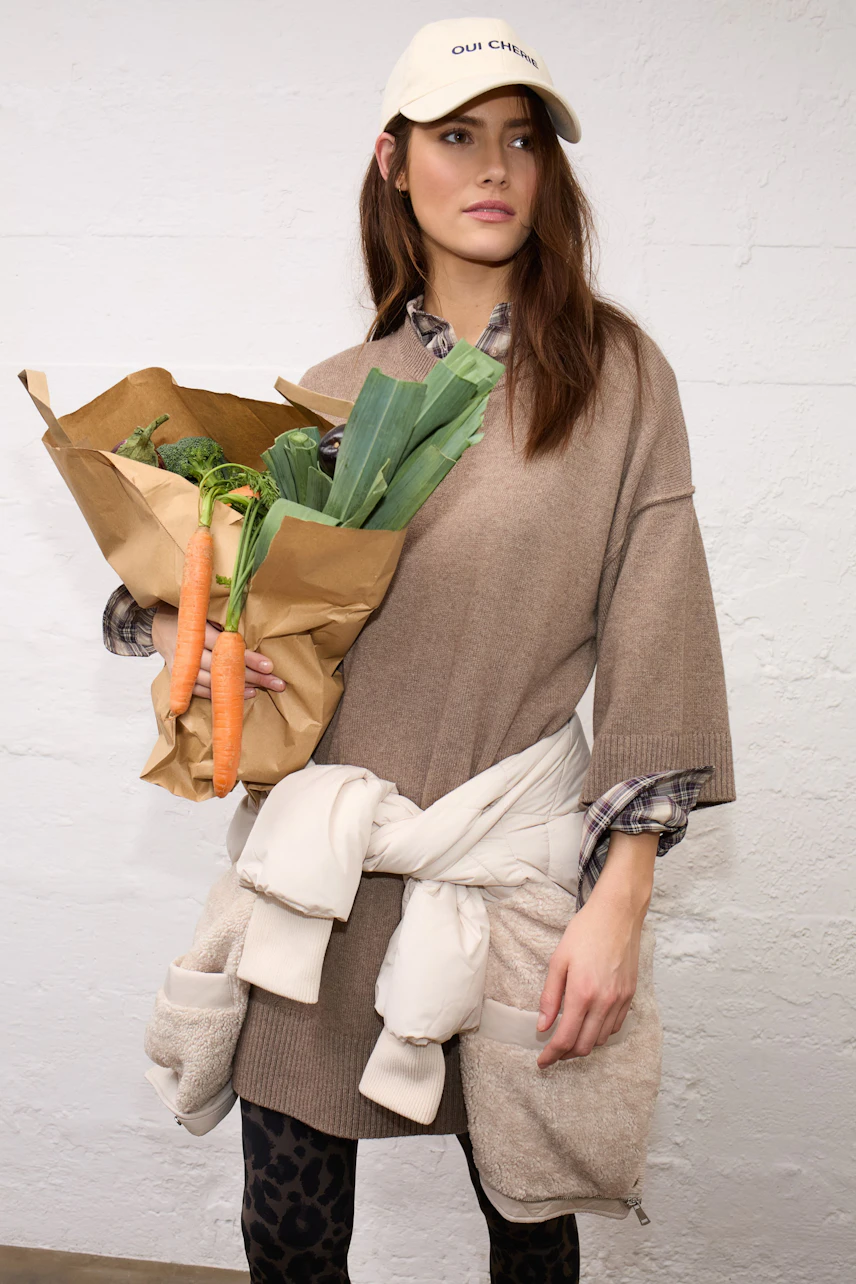 Casual women's hat: Woman in a beige cap, oversized sweater, and patterned leggings holding groceries.