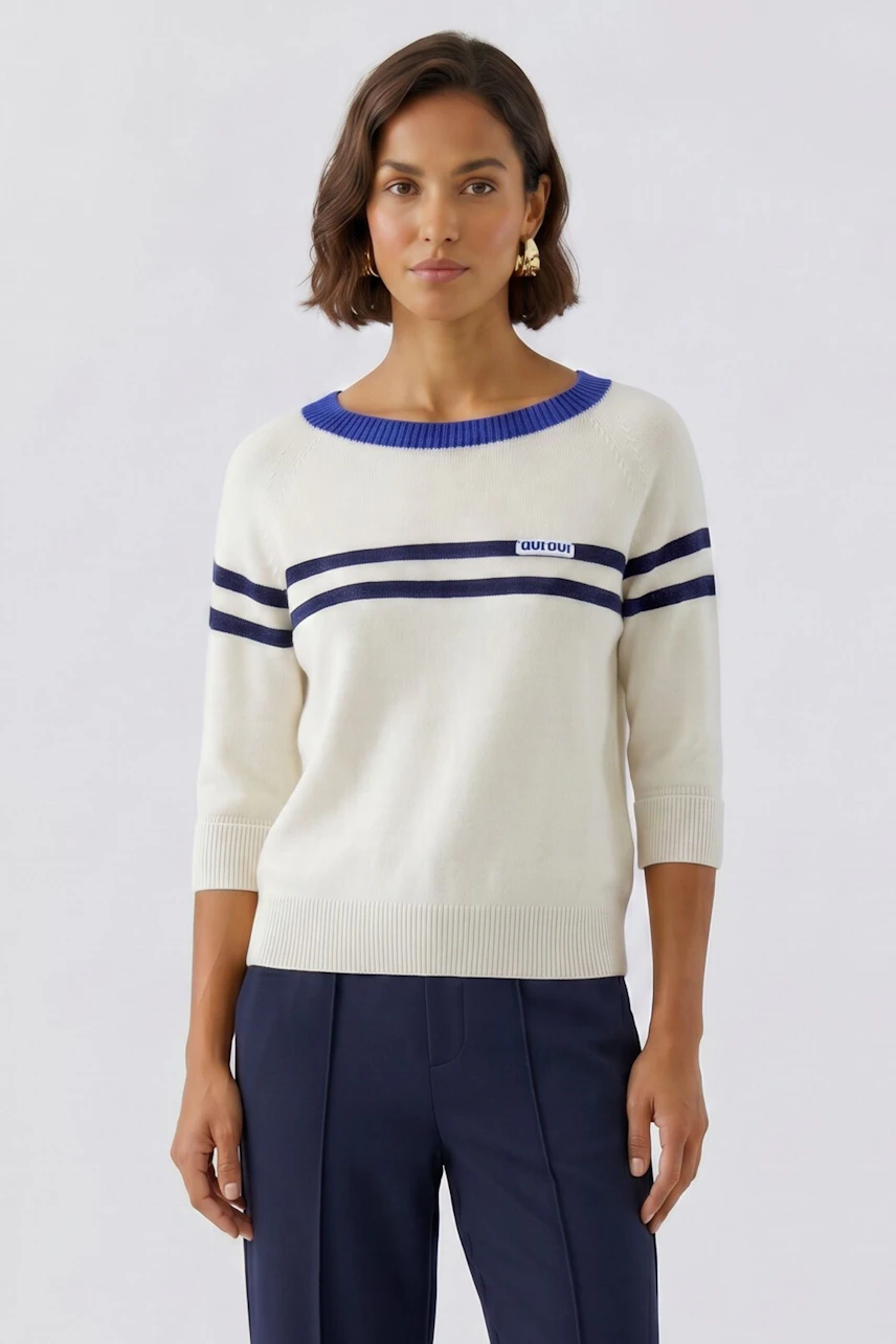 Woman in a white pullover with blue stripes and logo, paired with navy pants, standing confidently.