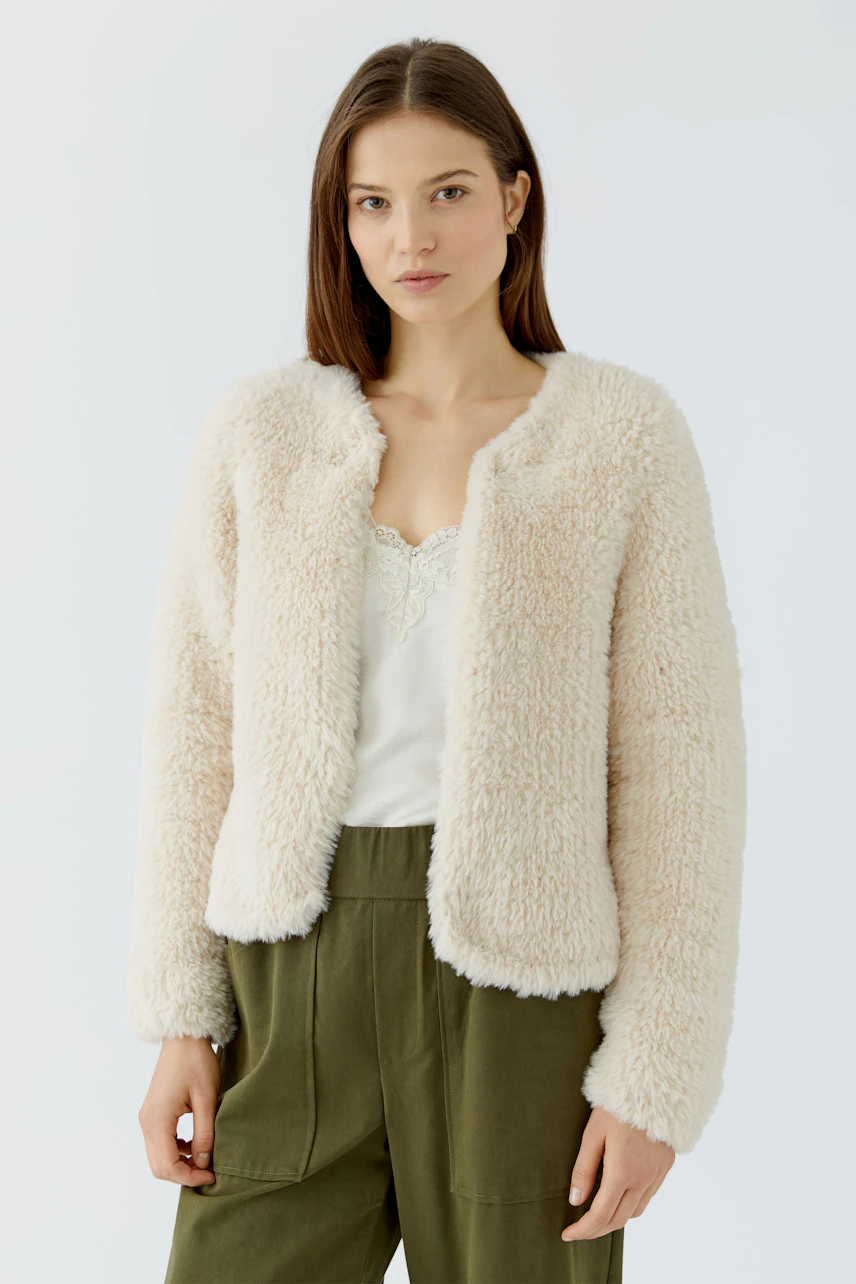 Frau in flauschiger Strickjacke: Cremefarbene Jacke über weißem Top und grüner Hose, lässiger Look.