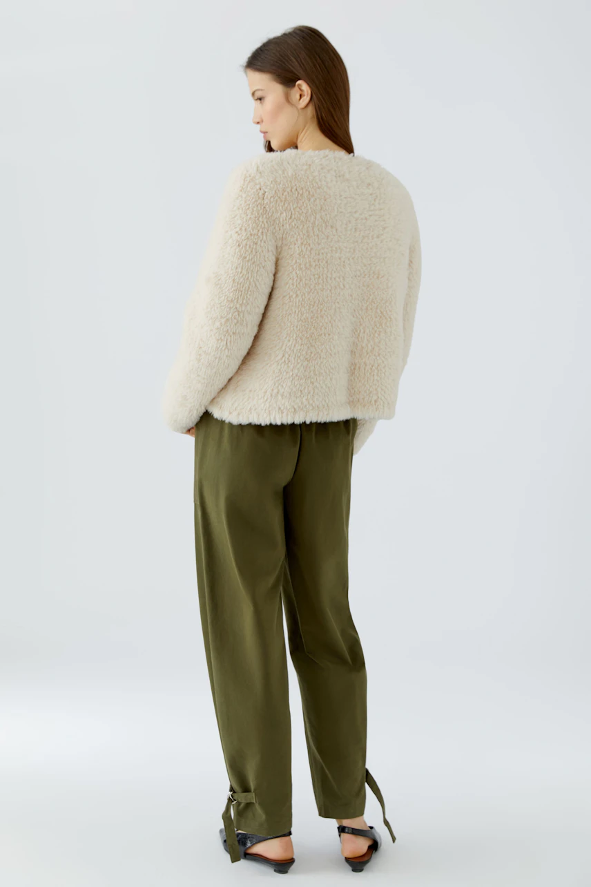 Frau in cremefarbener Strickjacke und olivgrüner Hose, seitlich stehend, mit Blick nach links.