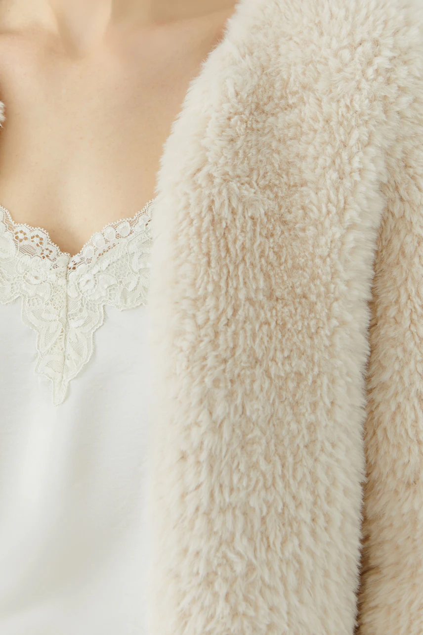 Flauschige Strickjacke: Frau in cremefarbener Strickjacke über weißem Spitzen-Top, weich und gemütlich.