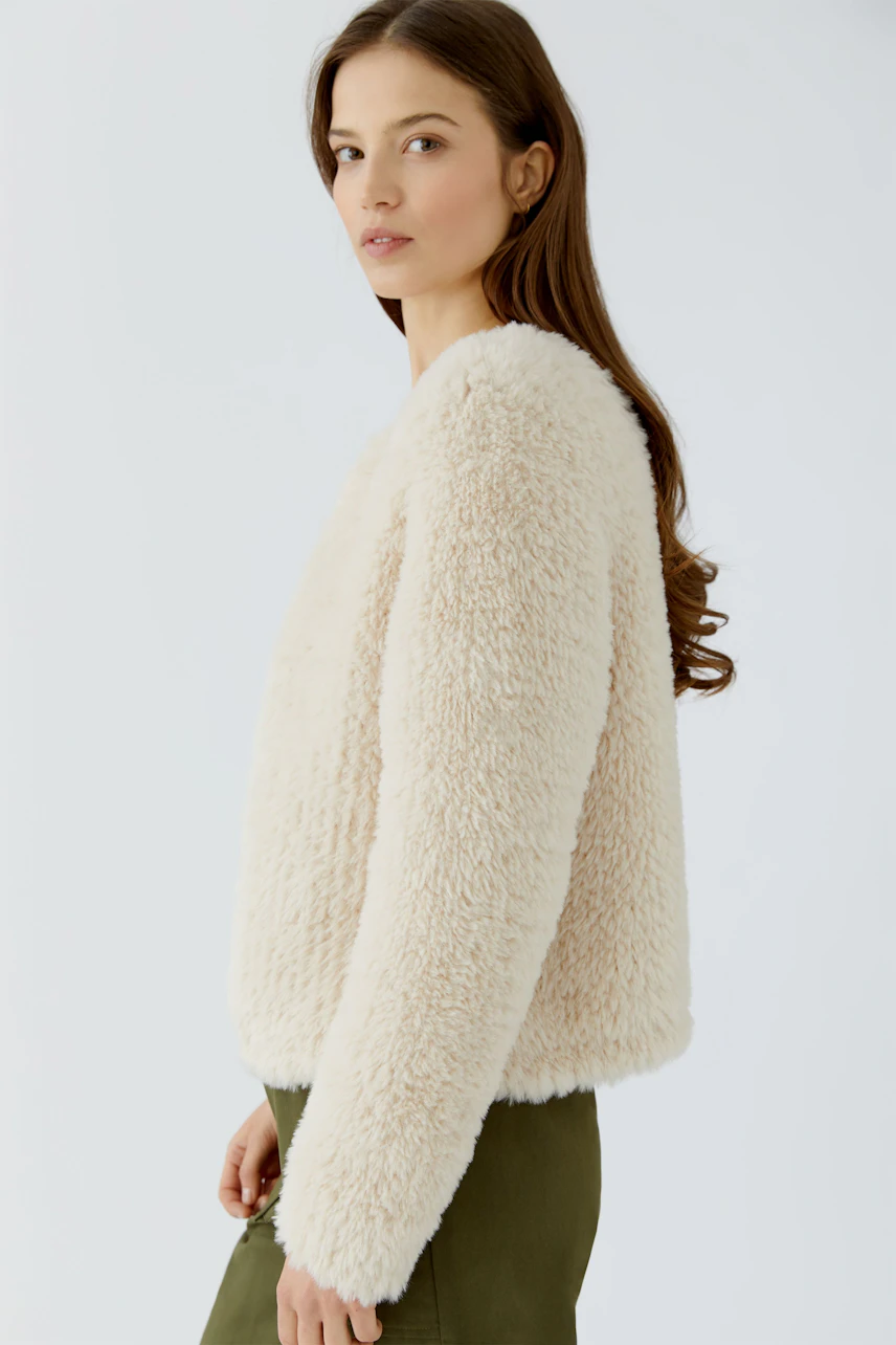 Frau in cremefarbener Strickjacke, seitlich stehend, kombiniert mit olivgrüner Hose.