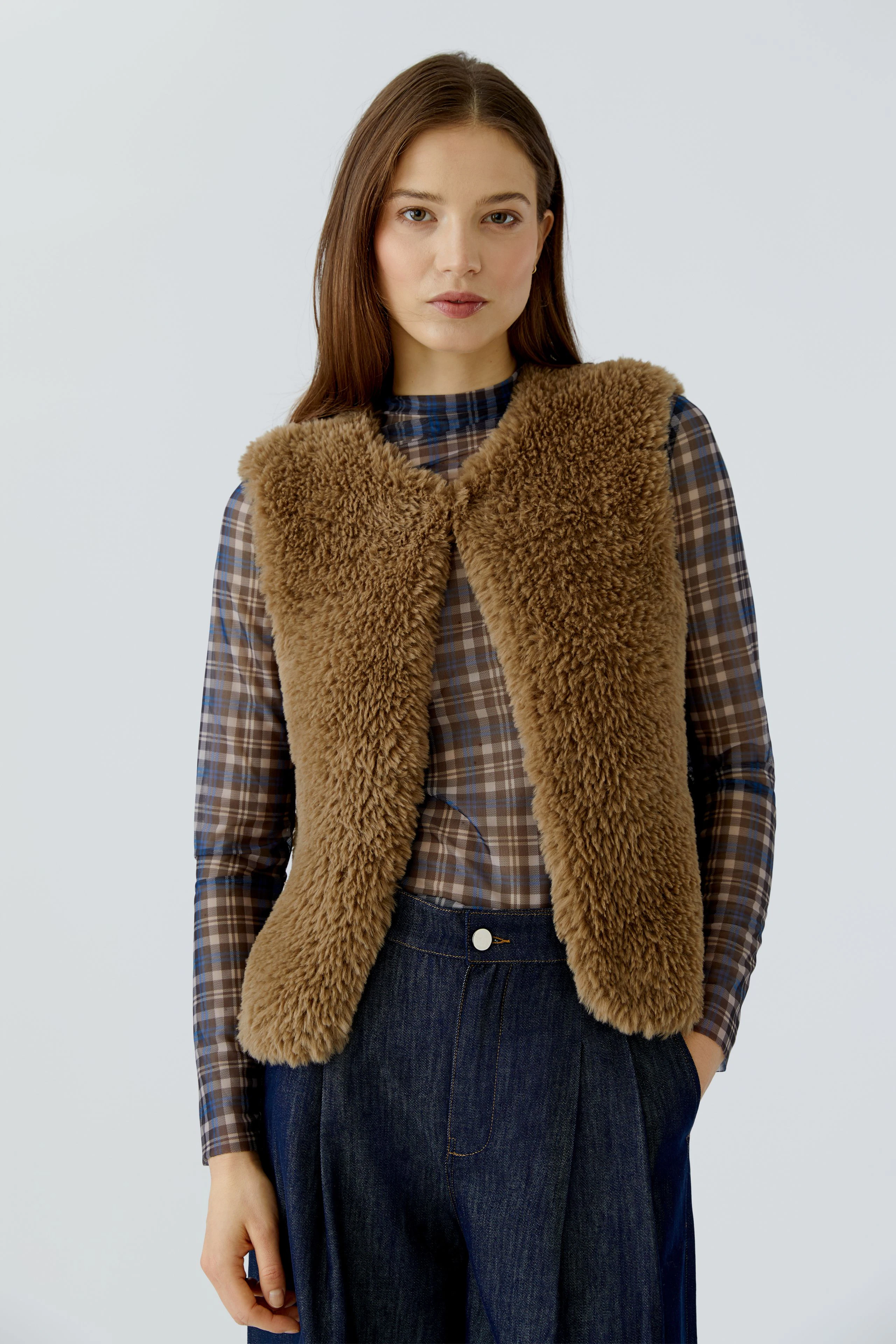Woman in a cozy brown knit vest over a plaid shirt, paired with dark denim jeans, looking confidently.
