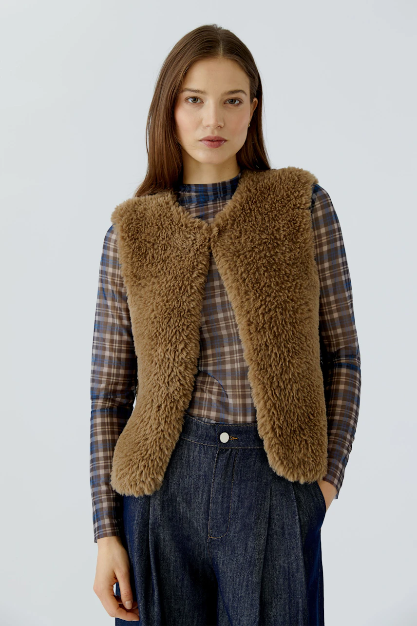 Woman in a cozy brown knit vest over a plaid shirt, paired with dark denim jeans, looking confidently.