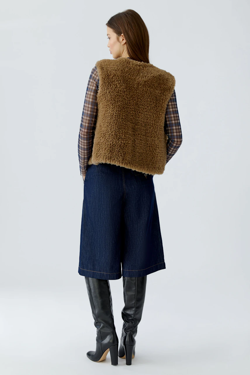 Woman in a cozy brown knit vest over plaid shirt, denim culottes, and knee-high boots, facing away.
