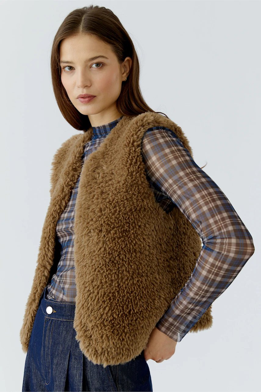Woman in a brown knit vest over a plaid shirt, paired with dark jeans, looking confidently to the side.