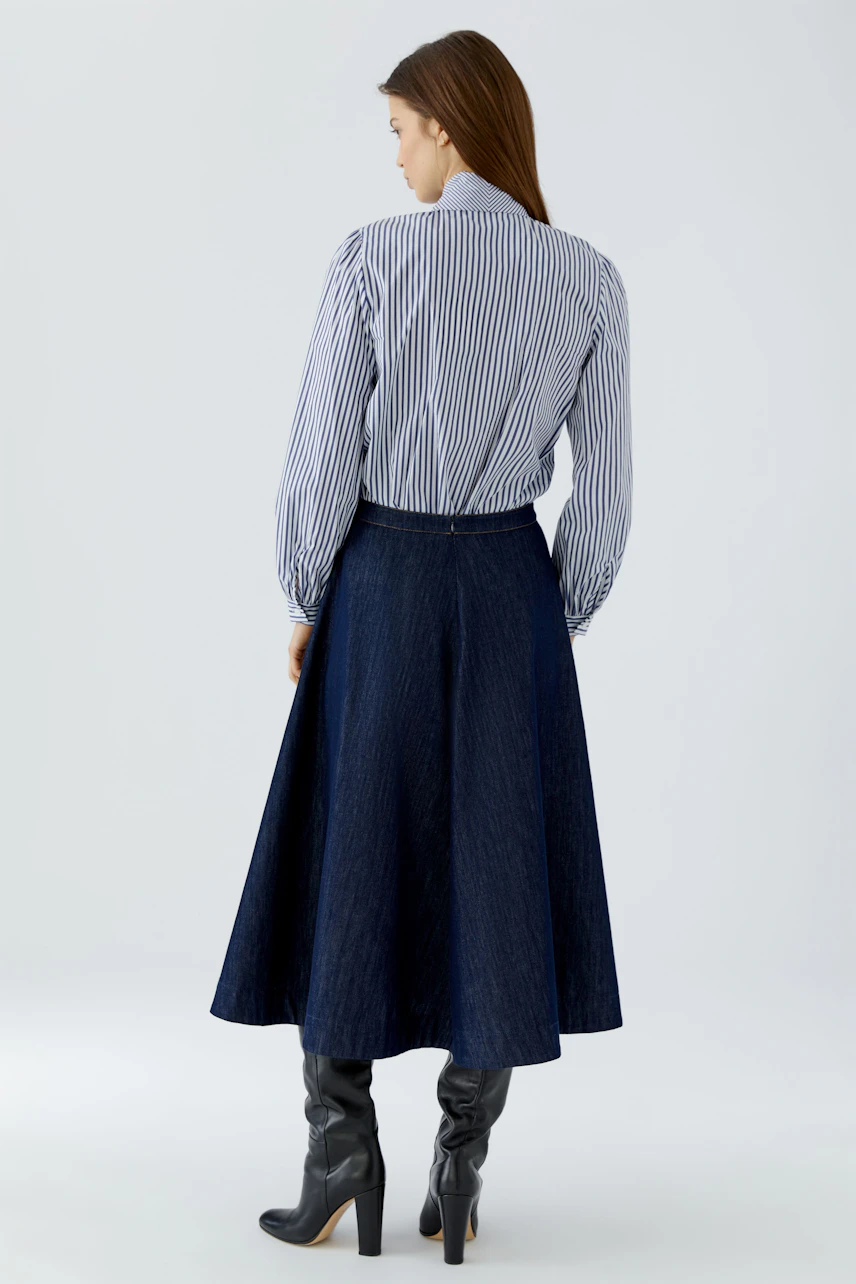 Frau in gestreifter Schluppenbluse und weitem Rock, blickt zur Seite. Lässiger Look mit Stiefeln.