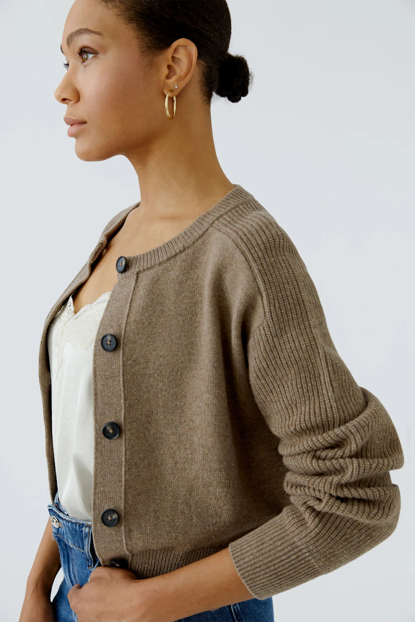 Strickjacke: Frau in brauner Strickjacke mit Knöpfen, weißem Top und blauer Jeans, seitlich blickend.