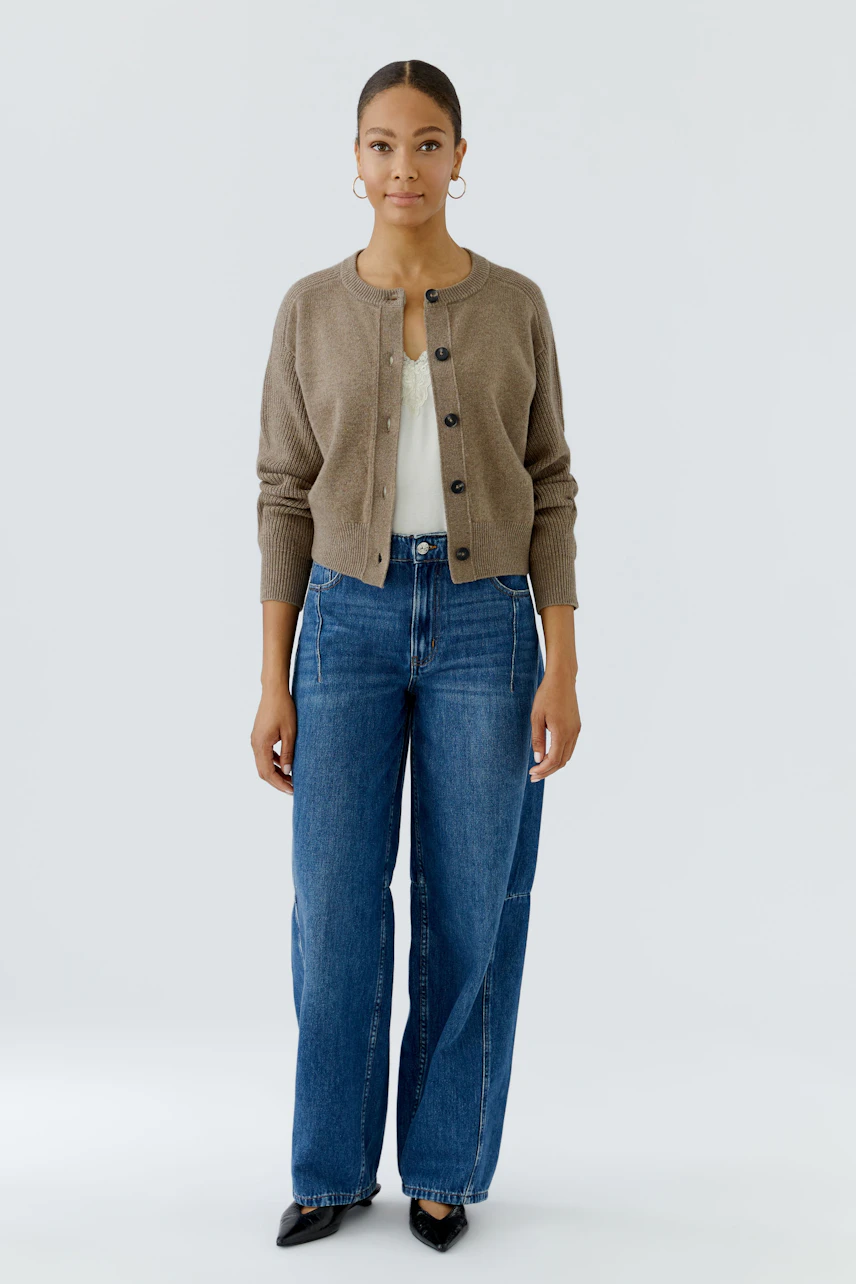 Casual cardigan: Woman in a brown knit cardigan, white top, and blue jeans, standing confidently.