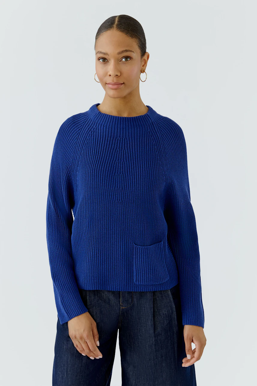 Woman in a blue pullover with a front pocket, paired with dark jeans, standing confidently.