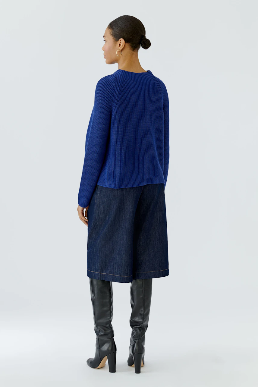 Woman in a blue pullover, denim culottes, and knee-high boots, viewed from the back.