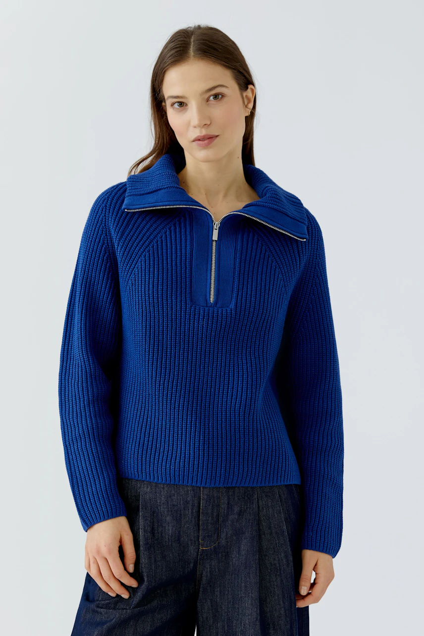 Woman in a blue ribbed pullover with a zip collar, paired with dark jeans, standing confidently.