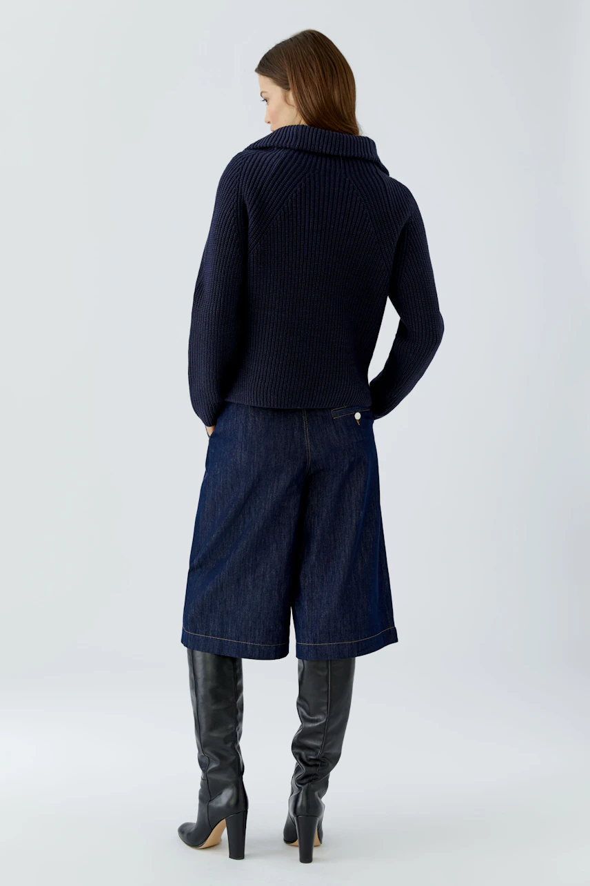 Woman in navy pullover, denim culottes, and knee-high boots, viewed from the back.