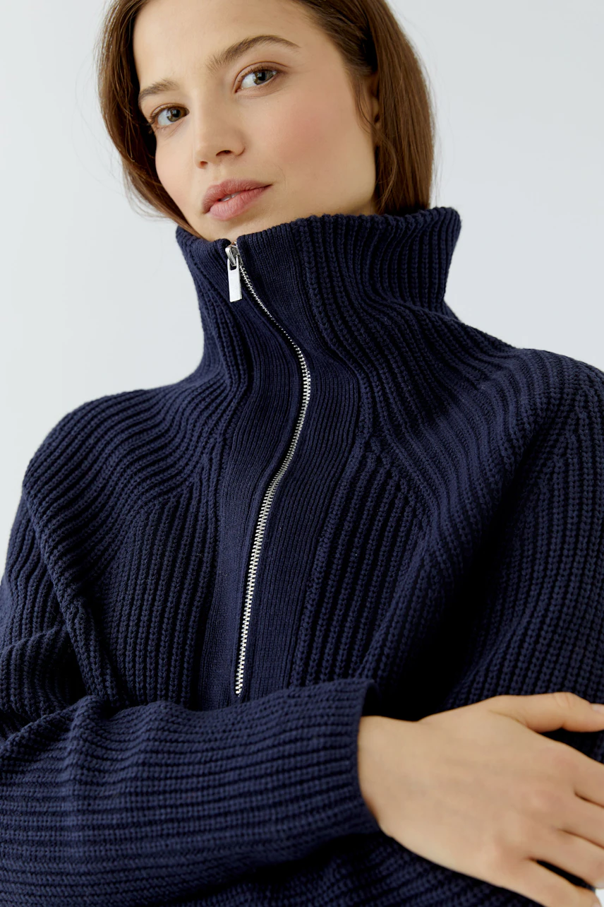 Woman in a navy ribbed pullover with a high collar and zipper, looking confidently at the camera.