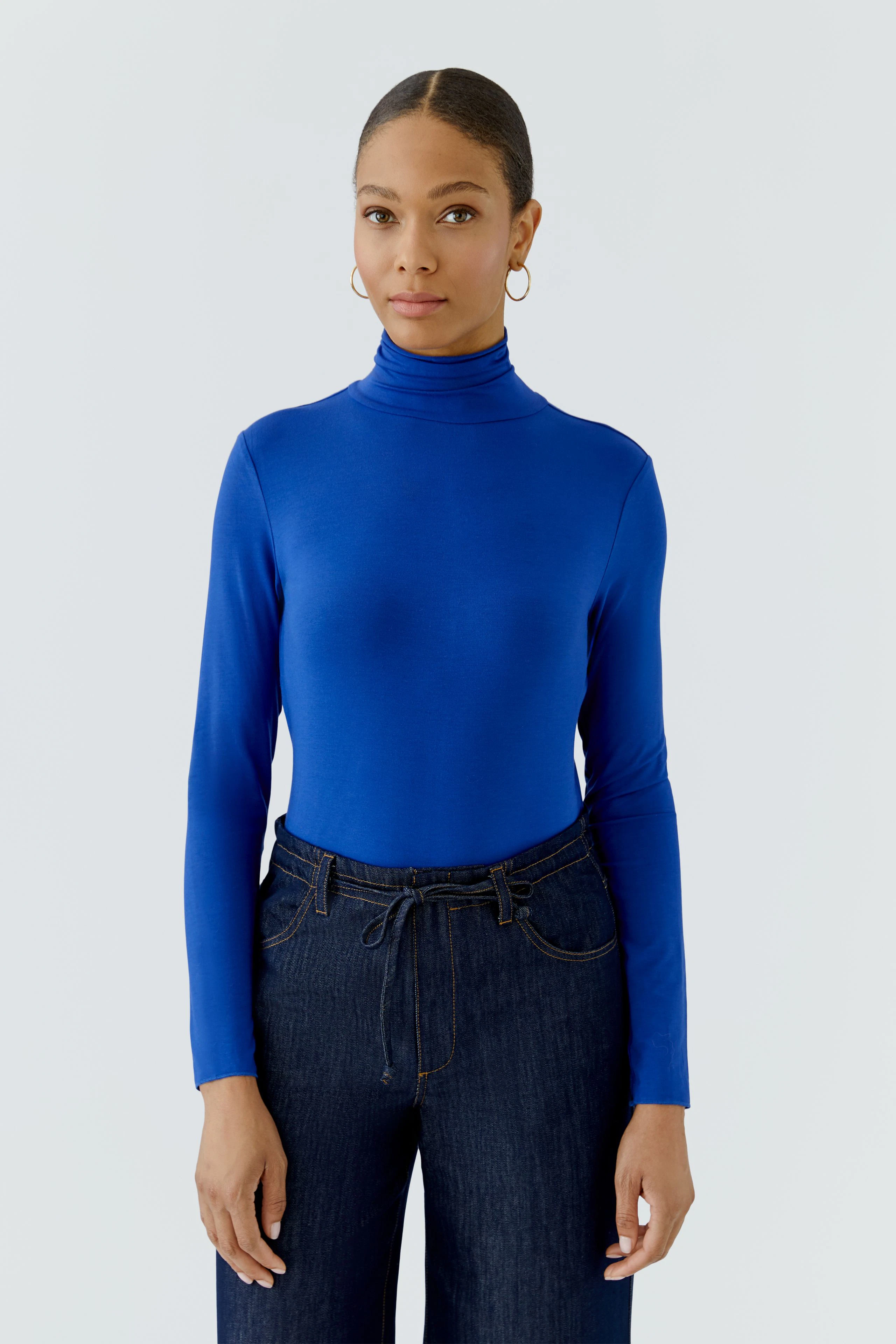 Woman in blue long-sleeve shirt with turtleneck, paired with dark jeans, standing confidently.