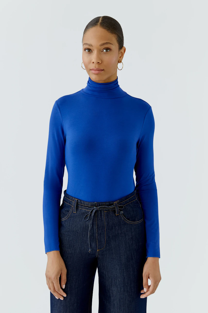 Woman in blue long-sleeve shirt with turtleneck, paired with dark jeans, standing confidently.